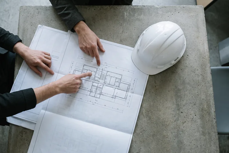 Two people point to a detail on unfolded drawings; a hard hat lies on the table.